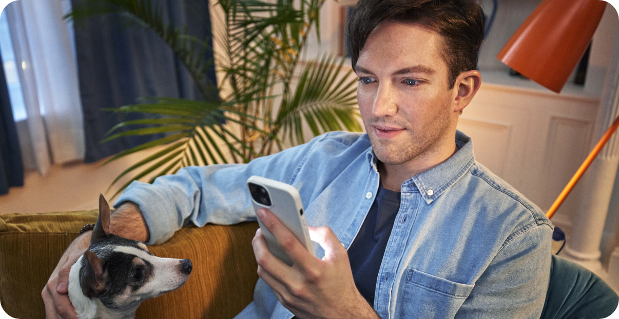 Man sitting on a couch with his dog, looking at his phone.